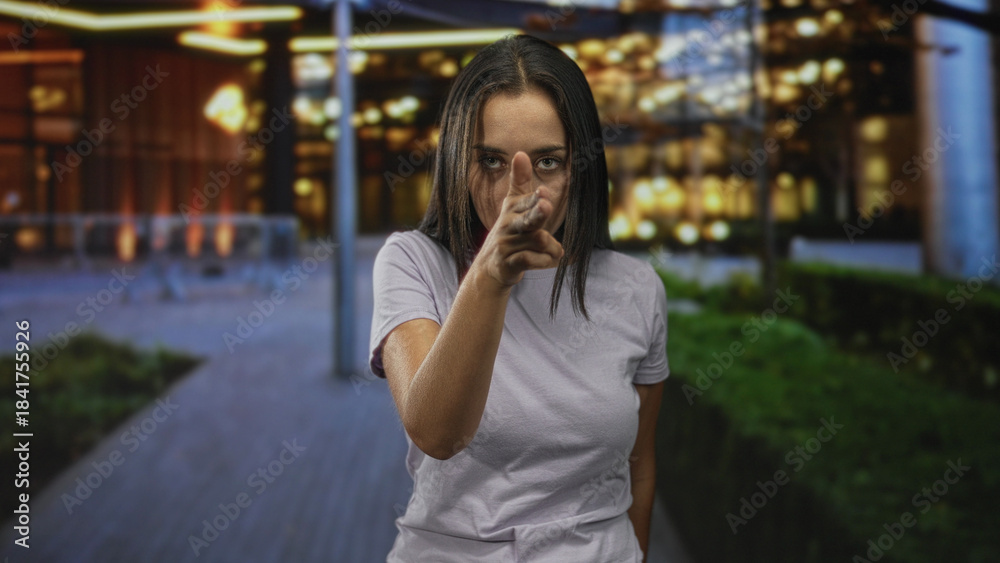 Fototapeta premium Woman pointing finger at camera and holding finger to lips for silence on street; secrecy seriousness.