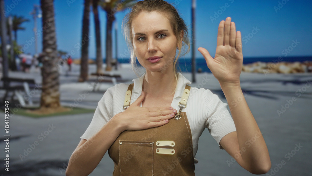 Φωτογραφία Young woman makes a pledge gesture wearing apron on sunny promenade by the seaside, capturing a moment of sincerity and tranquility outdoors with palm trees in the background