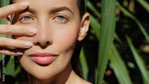 Luminous skincare model with green eyes showcases fresh, dewy skin as she frames her face with elegant fingers, glowing under natural sunlight against lush tropical leaves.