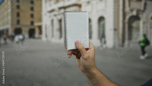 Billede på lærred Man holding notebook in outdoor city street scene with blurred buildings and people in the background during daytime, emphasizing urban life and public space