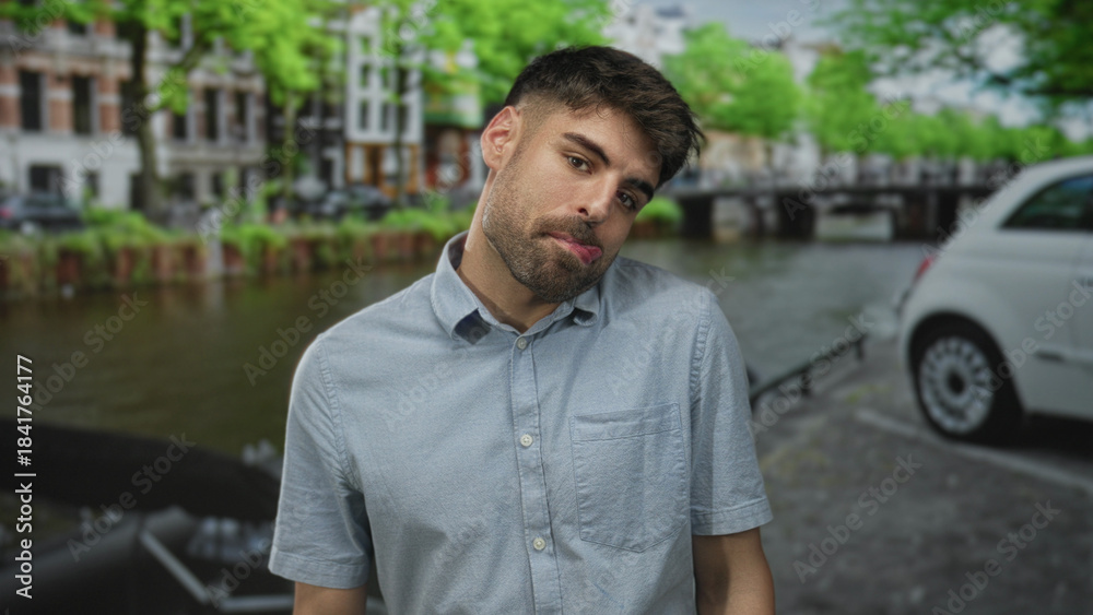 Fototapeta premium Man tilts head and pouts lips by canal on city street amid green tree canopy with buildings lining waterway; curiosity.