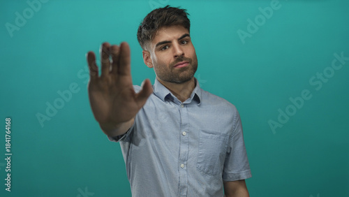 Young hispanic man pushes palm outward at camera in teal studio setting with serious expression; defiance.