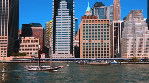 Beautiful ship with sails down goes by the blue waterscape. Low angle view on the stunning buildings of New York riverside at backdrop.