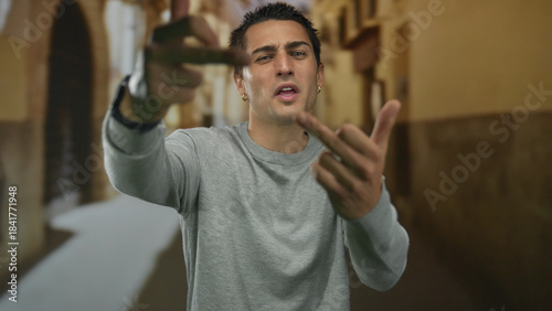 Young hispanic man shows middle finger gesture in narrow cobblestone old town street; anger defiance rejection.