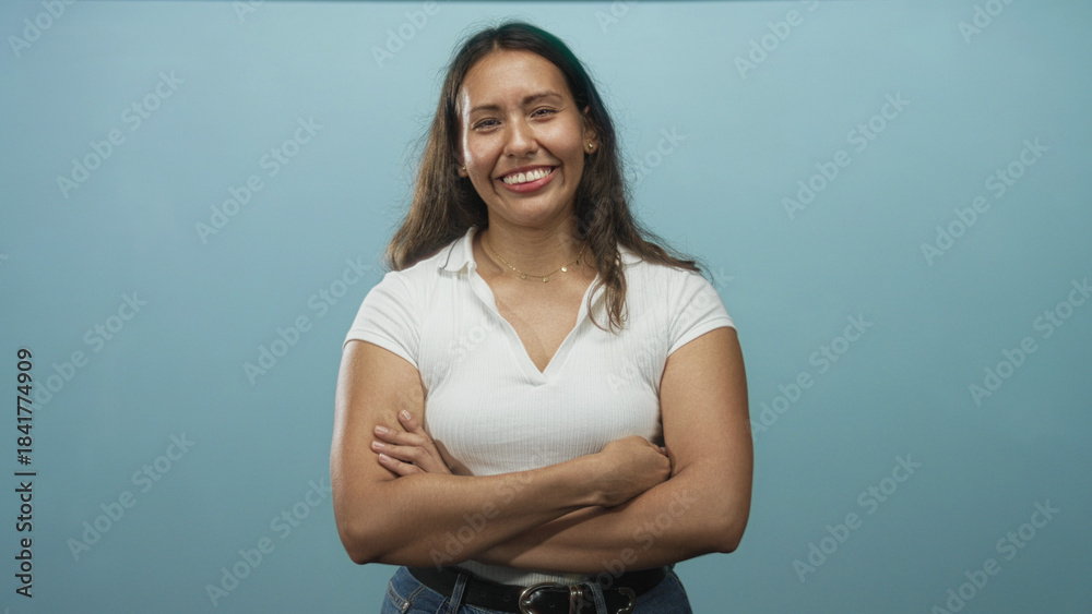 Obraz premium Woman with arms crossed smiling in blue studio, wearing a white top and jeans with belt, showing forearms and a delicate necklace; confident friendly approachable.
