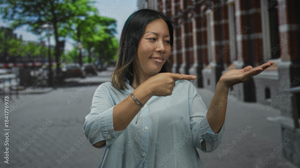 Fototapeta premium Woman points finger to open palm on city street while smiling and looking at hand; friendly invitation.