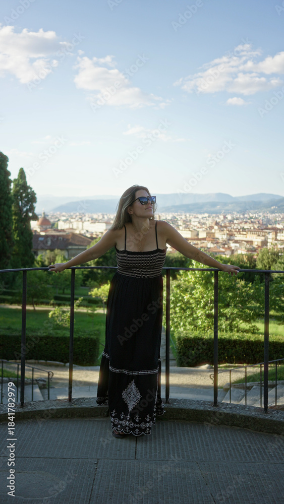 Fototapeta premium Woman standing with arms outstretched on viewpoint railing in florence wearing black sundress and sunglasses, bare feet visible; serenity.