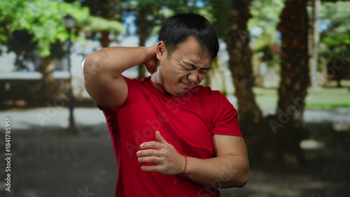 Young man in red shirt showing discomfort outdoors in a park setting with blurred trees in the background.