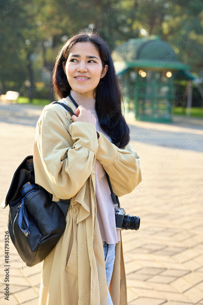 Naklejka premium Woman with camera and backpack stands in a sunny park, wearing a beige trench coat and soft scarf, ready for outdoor photography, exploration, and casual storytelling; natural light highlights her