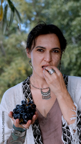 A beautiful brunette in a bohemian style relaxes outdoors in summer against a backdrop of a green forest, holding a large bunch of juicy black grapes