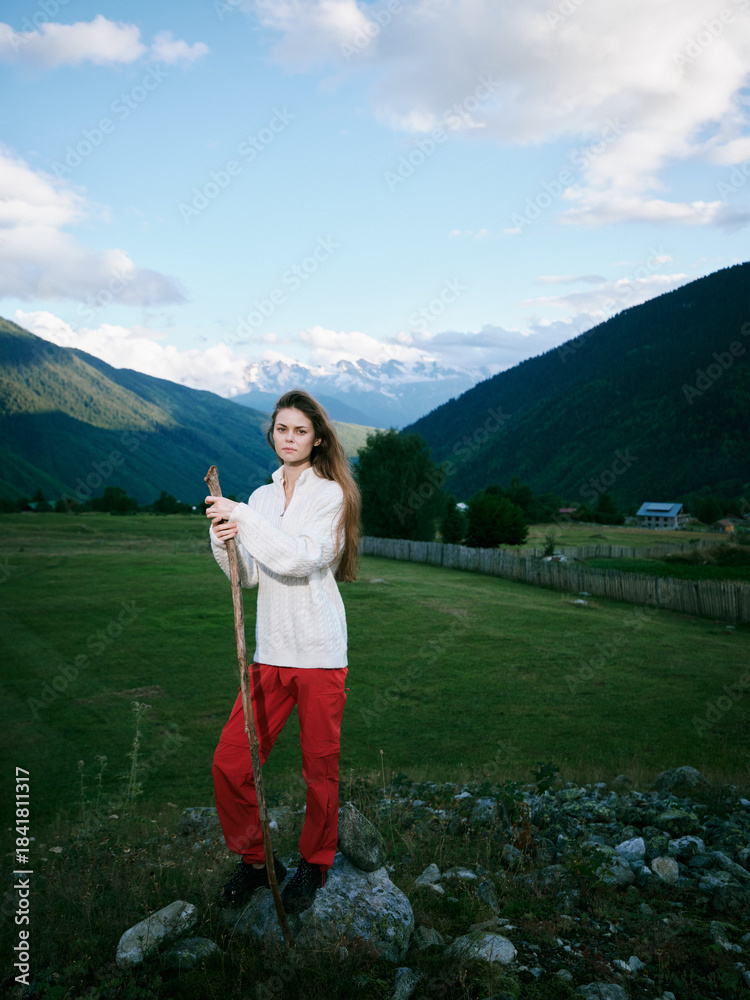 Naklejka premium Mountain field outdoor scene with a woman in red pants holding a staff, valley views, clear sky and rustic fence, capturing nature and exploration in wide rural landscape
