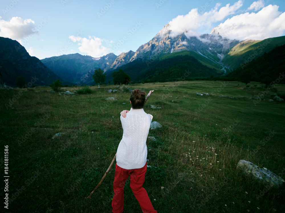 Naklejka premium Back view of a hiker in a white top and red pants standing in a wide meadow with rocks, pointing toward distant mountains under a bright blue sky and clouds