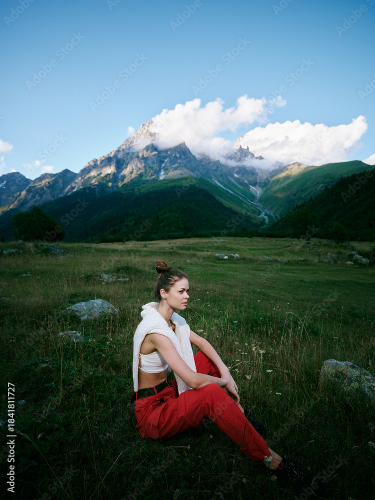 Naklejka premium Woman sits in a green meadow at the foot of towering mountains, wearing red pants and a white top, gazing into the distance under a blue sky with fluffy clouds during outdoor travel
