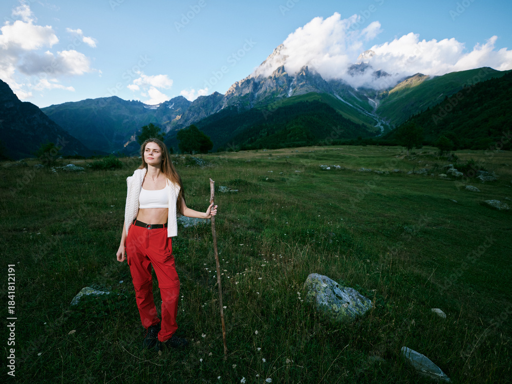Naklejka premium Woman standing in a mountain meadow with a walking stick, wearing red pants and a white top, expansive landscape with distant peaks and open outdoors