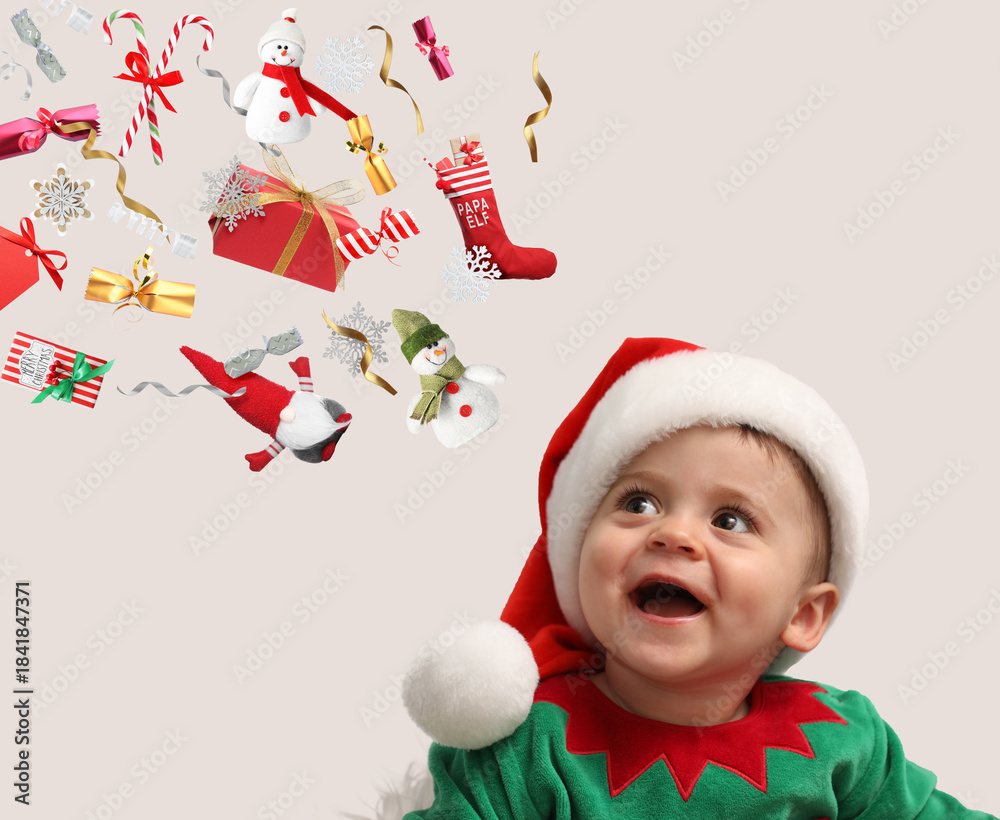Fototapeta premium Cute little baby in elf costume looking at flying festive stuff on light grey background. Christmas celebration