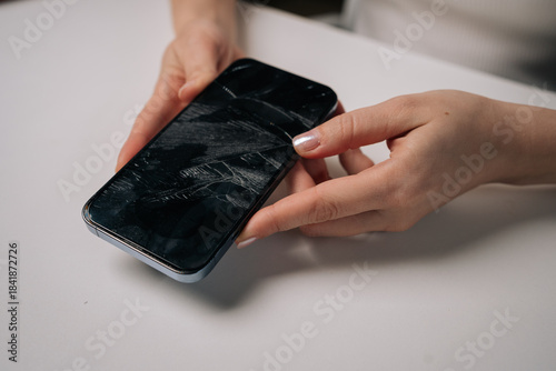Close-up of woman removing broken tempered glass screen protector from smartphone, revealing cracked protection film on white table. Concept of mobile phone maintenance and service.