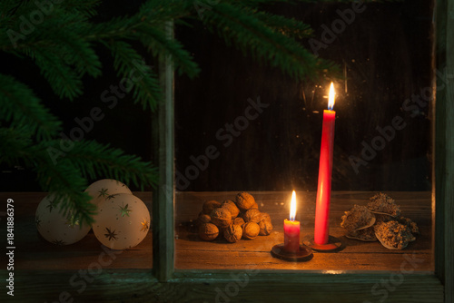 Through the old window in the darkness, two red candles glow. Their warm light illuminates Christmas ornaments, walnuts, and homemade macaroons. A sprig of pine frames the festive scene.