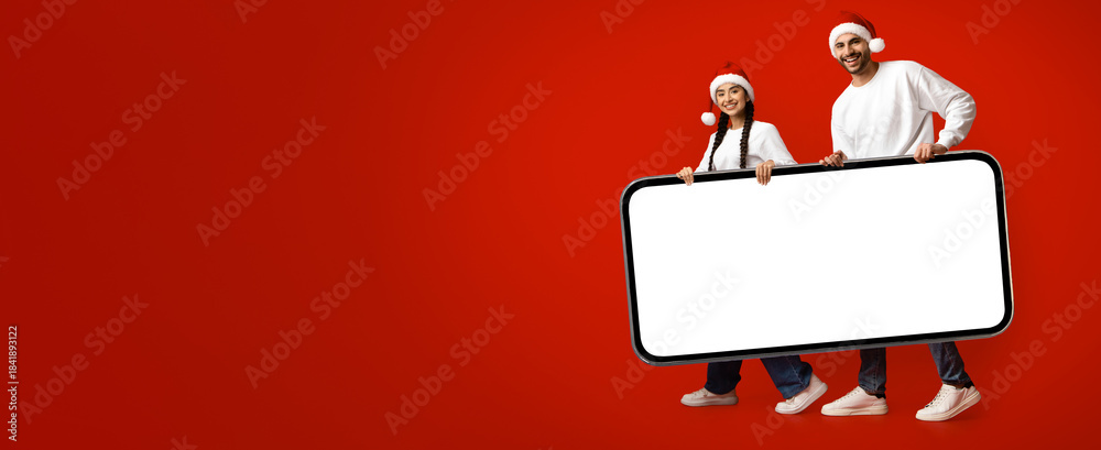 Obraz premium A man and woman, both wearing Santa hats, smile while holding a large blank sign in front of a vibrant red backdrop. The scene captures a festive holiday spirit.