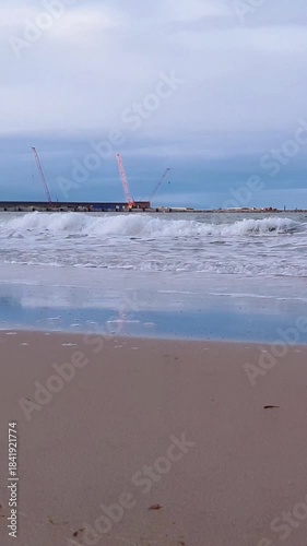 Low angle vertical shot stormy waves breaking near coastline on desolate sand beach on Baltic sea shore and seaport quay on background under cloudy sky at overcast day. No people tranquil seascape.