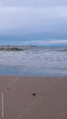 Vertical panoramic shot of stormy coastal surf water waves rolling on empty sand beach on Baltic sea coast and seaport quayside on background at cloudy autumn day. No people desolate seascape.