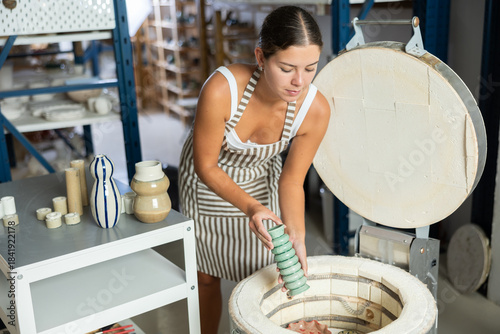 Young potter girl takes out finished ceramic dishes, various mugs and a vase from the oven