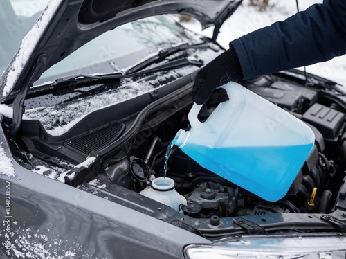 A person pours blue antifreeze windshield washer fluid into a car reservoir during a snowy winter day.