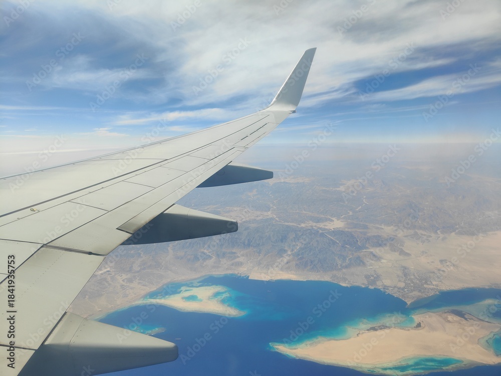 Obraz premium Aerial View of Airplane Wing Over Coastline and Sea, Red Sea, Egypt - A high-resolution aerial photograph taken from an airplane window, showing the aircraft wing above a deep blue sea with a desert c