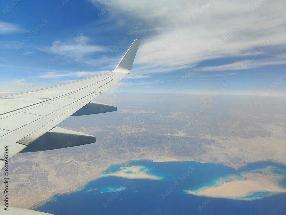 Obraz premium Aerial View of Airplane Wing Over Coastline and Sea, Red Sea, Egypt - A high-resolution aerial photograph taken from an airplane window, showing the aircraft wing above a deep blue sea with a desert c
