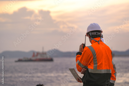 Photography Two engineers wearing safety uniforms and hard hats communicate using a radio while reviewing data on a laptop by the sea during sunset