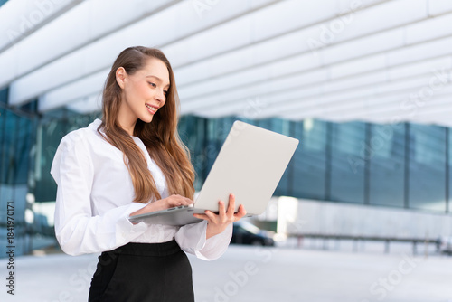 Young professional woman in white shirt standing outside glass building using laptop representing business, remote work, technology and urban corporate success Free Download Young professional woman in white shirt standing outside glass building using laptop representing business, remote work, technology and urban corporate success - GFXLoad