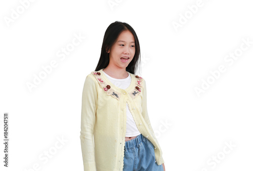 Cheerful young Asian girl in light yellow cardigan and denim jeans standing and looking to the side with an open mouth, possibly speaking or laughing, isolated on white background.