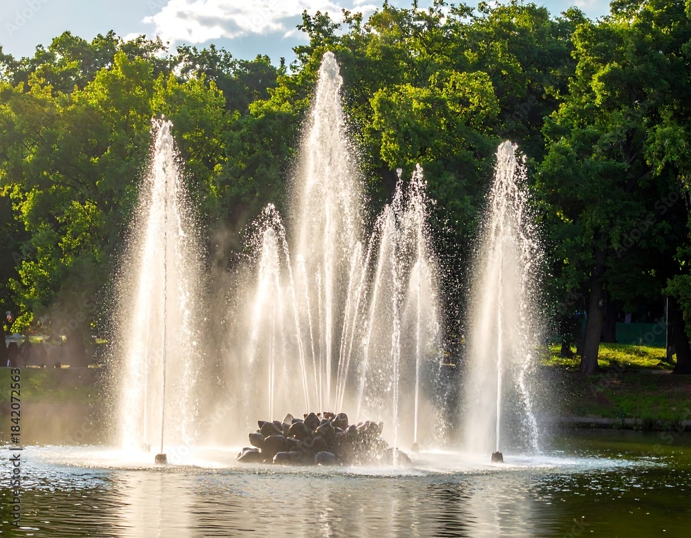 Naklejka premium Fountains in a pond, illuminated by sunlight, with green trees in the background