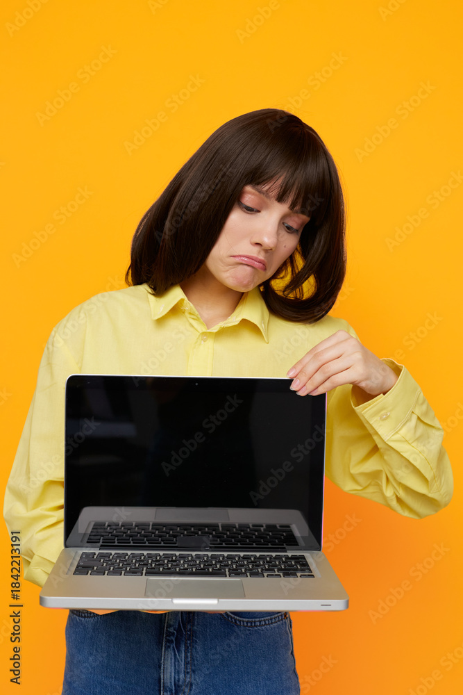 Fototapeta premium A young woman in a yellow shirt holds an open laptop, gazing downward with a pout as bright orange backdrop highlights a moment of tech frustration or focus.