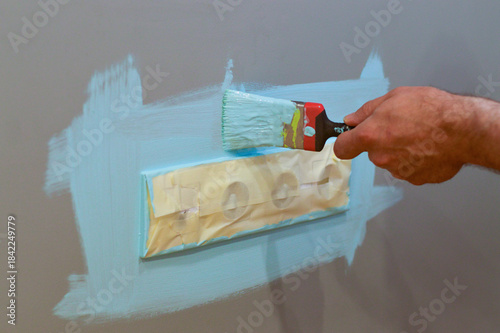 Home interior update involving painting around an electrical outlet with a brush, employing masking tape for protection, as part of a DIY project to maintain a tidy and safe space.