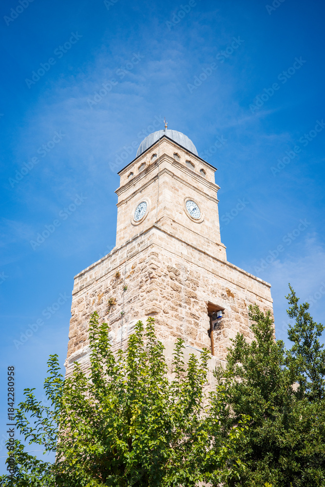 Obraz premium Low-angle view of a historic stone clock tower rising above green trees under a clear blue sky in Turkey. The timeless architecture, weathered limestone texture, and classic clock tower