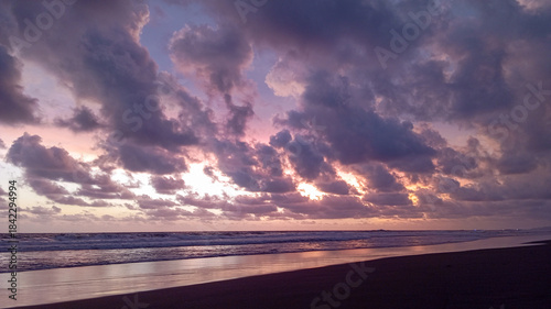 A breathtaking sunset over a peaceful beach featuring dramatic purple clouds, rolling ocean waves, and a soft golden horizon glow