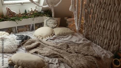 Warm evening in interior bedroom with bed and linens. Christmas decor or Newyear. Cozy space with plaids and pillows, shelf with branches fir with illumination garland glowing lights at home for relax