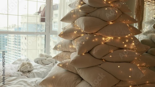 Soft Christmas tree made of many cushion with illumination garland glowing lights. Cozy and warm space with pillows at home. Modern festive interior design living room. 