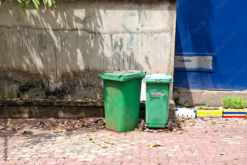 Obraz premium Two Green Outdoor Trash Bins Stand Next To A Textured Wall And Blue Structure