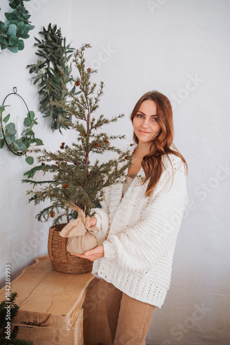 Young woman holding small potted evergreen tree indoors, warm cozy home decoration