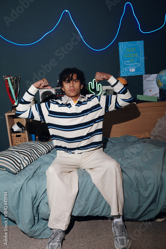 Teenage Black boy sitting on bed flexing arms and showing biceps, looking confidently at camera in modern bedroom with neon lights and coding themed decorations visible