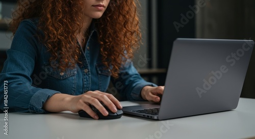 Woman with red curly hair in denim shirt working on laptop, using mouse at desk. Freelancer at home.