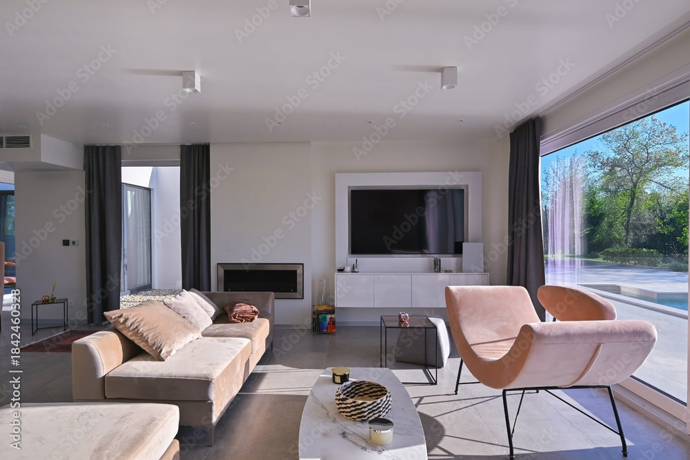 Obraz premium Modern luxury living room interior at sunset. Open-plan space featuring beige sofas, marble dining table, large glass windows with sun flare, and view of a patio. Stylish contemporary home design