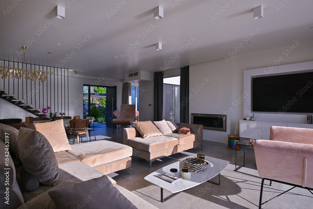 Obraz premium Modern luxury living room interior at sunset. Open-plan space featuring beige sofas, marble dining table, large glass windows with sun flare, and view of a patio. Stylish contemporary home design