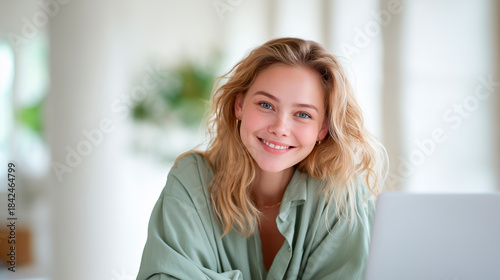 Smiling young woman in a sage shirt looks confidently into the camera while working on her laptop in a bright, cozy interior.