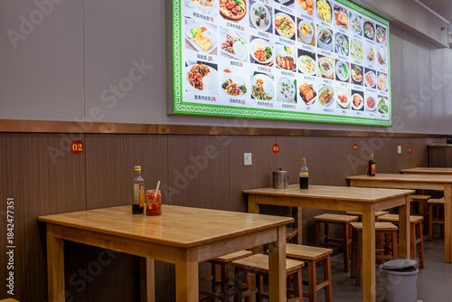Fototapeta Naklejka Na Ścianę i Meble -  Interior of small Chinese cafe in Sanya, Hainan, with simple wooden tables and stools beneath large illuminated menu board showing photos of dishes. Quiet, everyday local eatery atmosphere.