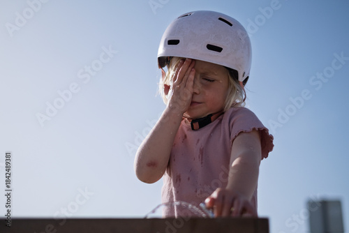 A little girl wearing a helmet drinks water from a public fountain and wipes her face. 