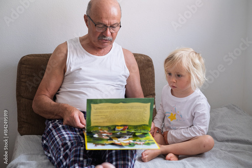 A grandfather is reading a book to his granddaughter while sitting together on a sofa at home. 