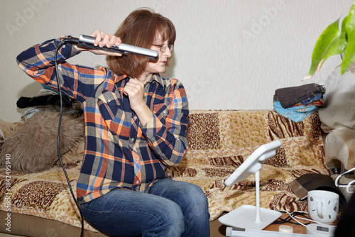 Middle-aged woman straightening her hair at home. Woman using hair straightener and focusing on hair styling. Haircare and hair styling concept
