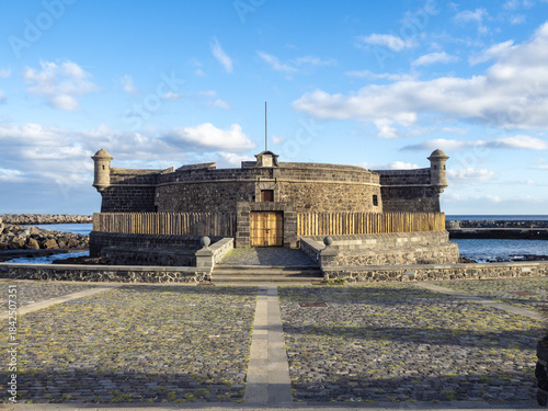View of the Castillo de San Juan Bautista, a historic stone fortress with a wooden door and a pole, under a clear blue sky, Tenerife, Canary Islands, Spain.